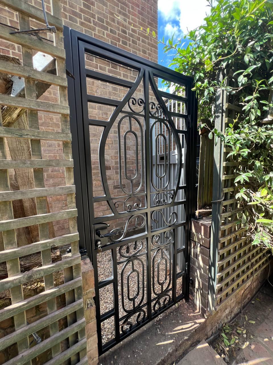 Full view of the decorative black metal garden gate installed between brickwork and timber garden screening.