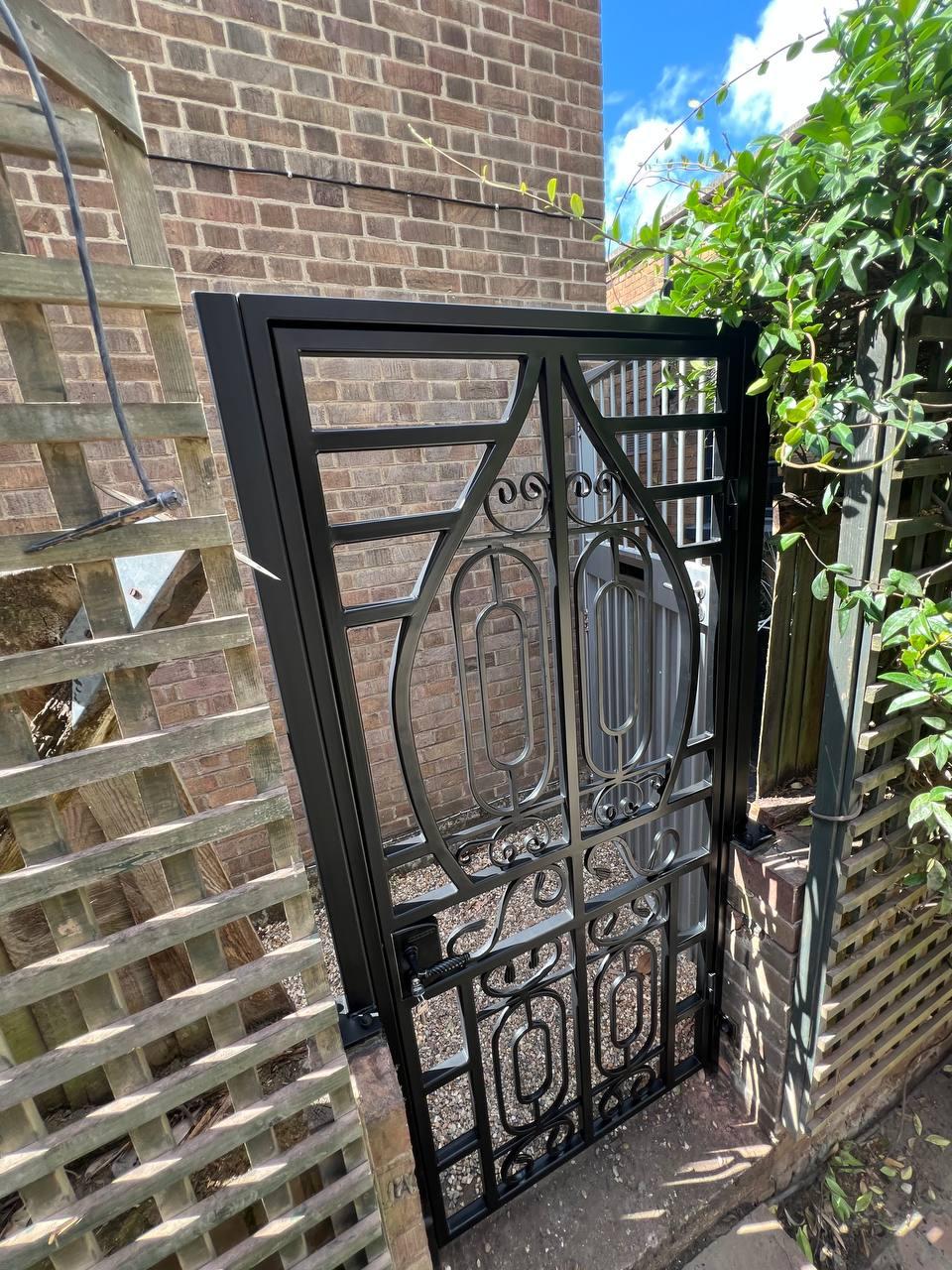 Decorative black metal garden gate with rectangular framing, elongated oval motifs and curled scrollwork beside a brick property.